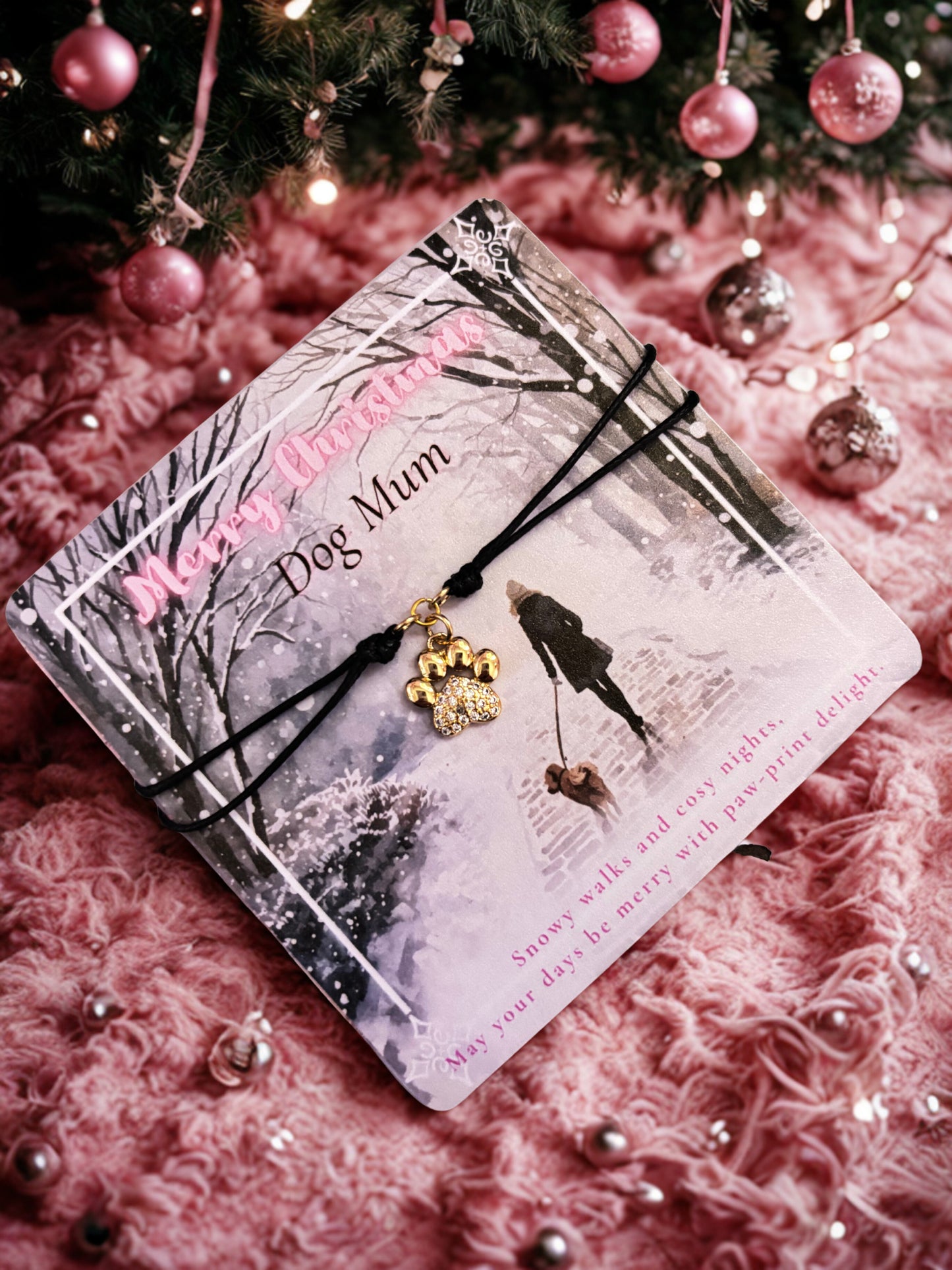 Wintery card with 'Merry Christmas Dog Mum' with a paw-print charm bracelet on a pink textured surface with Christmas decorations.