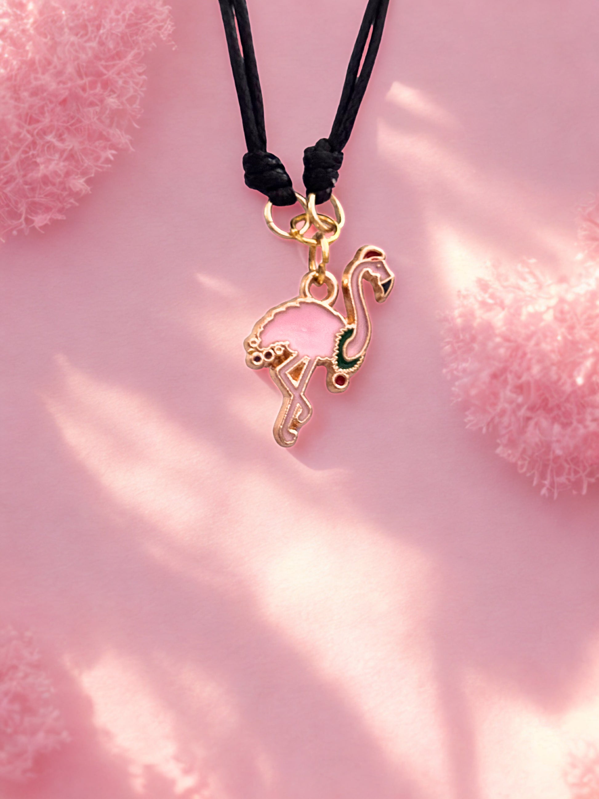 Gold flamingo charm on a black cord against a pink background