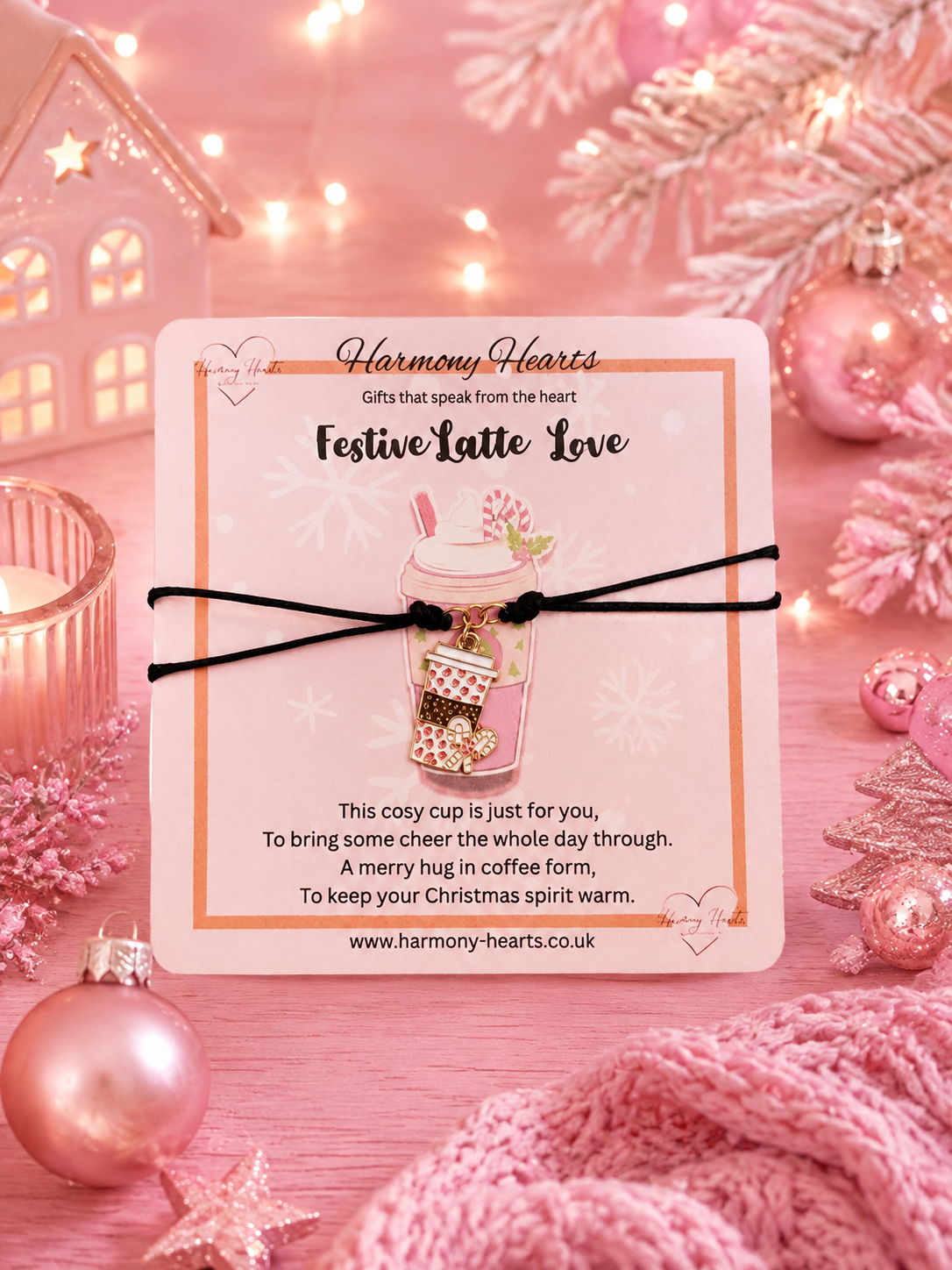 A pink Christmas theme card with a black cord braclet and coffee cup charm, styled on a pink Christmas-themed background with ornaments, lights, and a knit blanket. The card is branded Harmony Hearts and reads 'Festive Latte Love'.