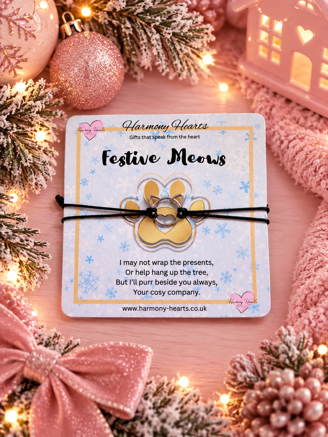 A festive paw print charm bracelet on a black cord, presented on a branded 'Harmony Hearts' gift card. The card is styled in a cozy Christmas setting with pink ornaments, pine branches, and warm fairy lights.
