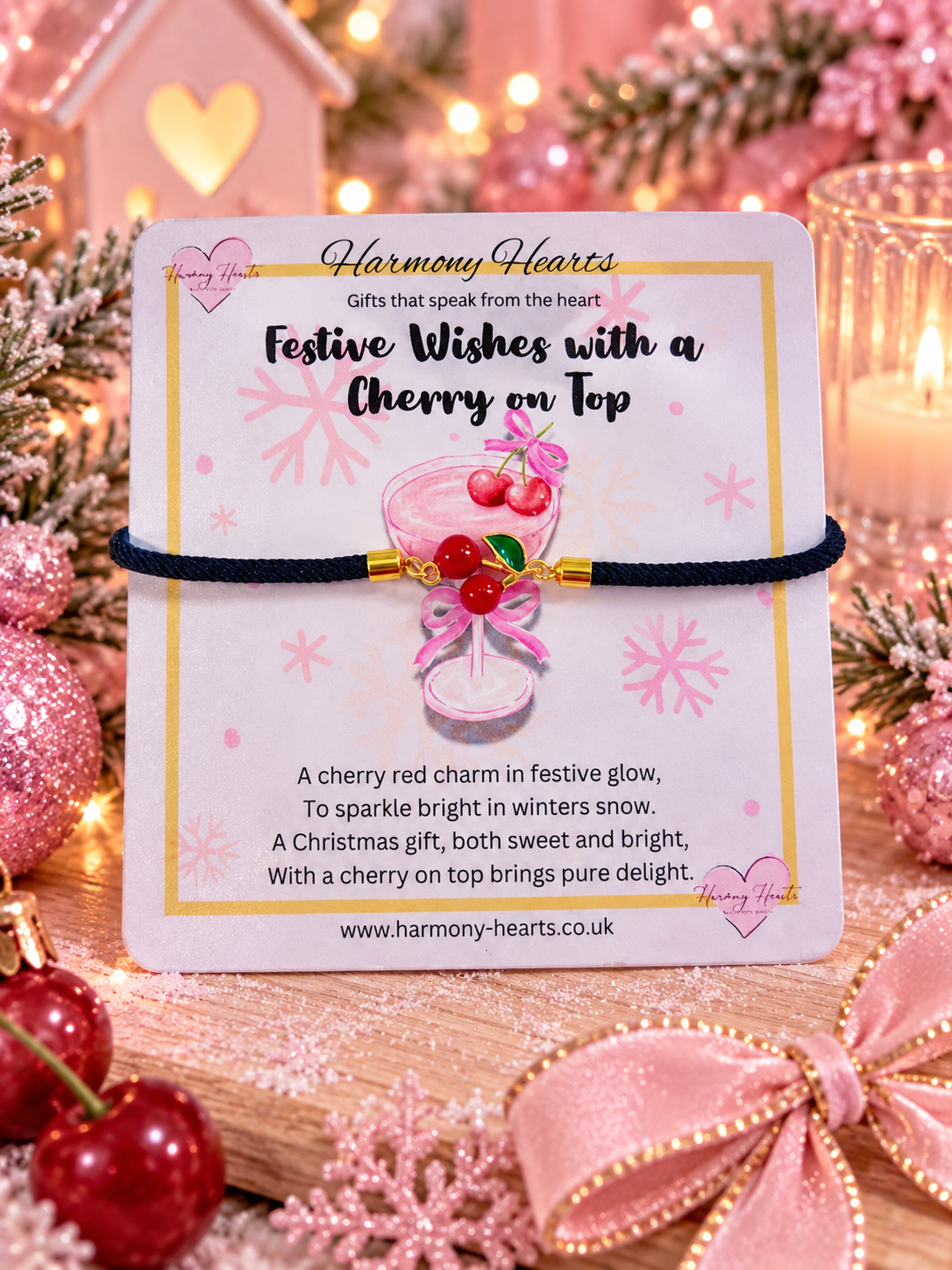 A cherry-themed charm bracelet on a black cord, mounted on a white 'Harmony Hearts' gift card. The card is styled on a wooden surface with festive Christmas decorations, including red cherries, pink glitter, a pink bow, and warm bokeh lights in the background.