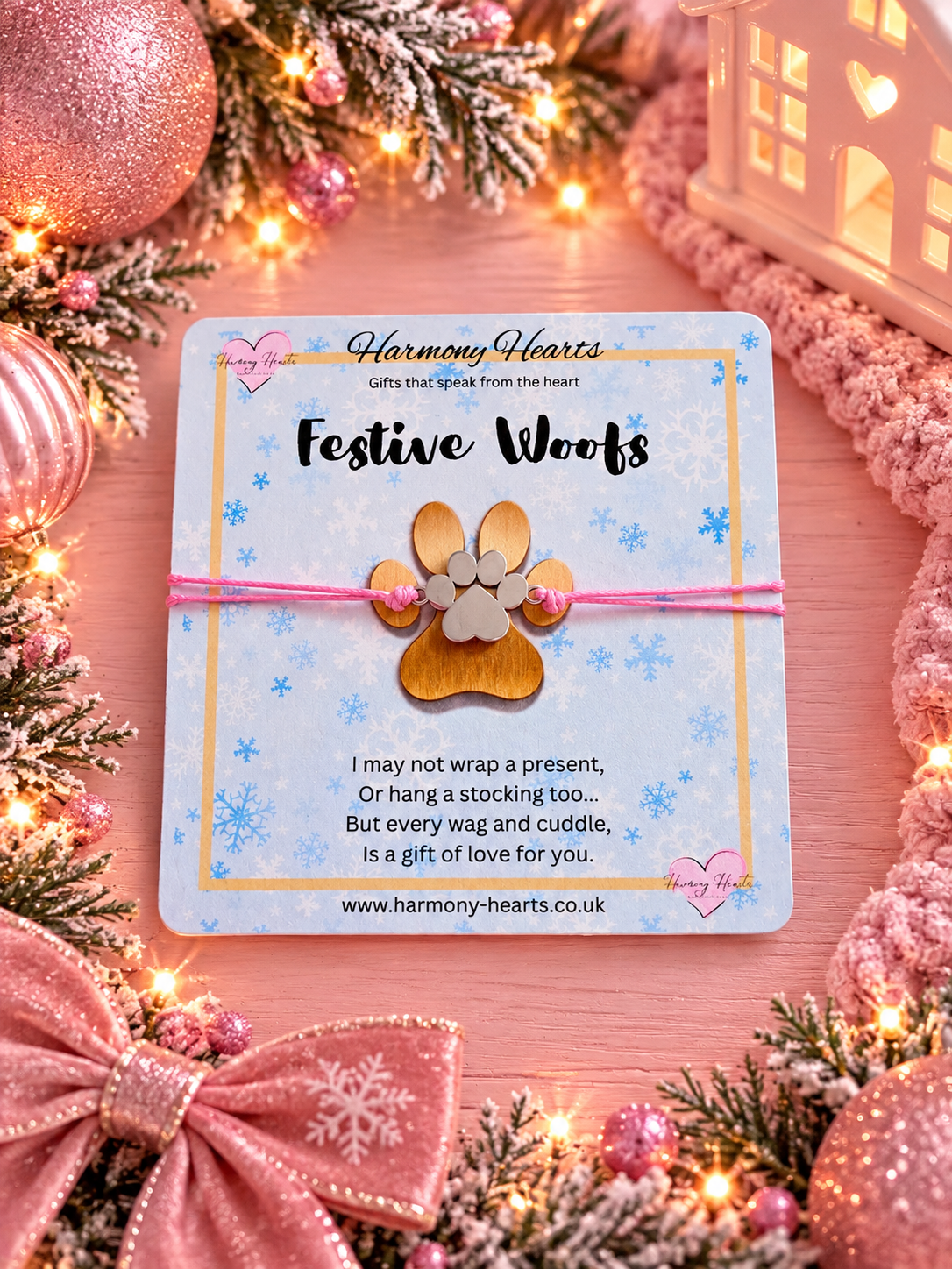 A handmade bracelet with a gold-colored paw print charm on a pink cord, presented on a branded 'Harmony Hearts' gift card. The card is styled in a festive Christmas setting with pink ribbons, ornaments, pine branches, and warm fairy lights.