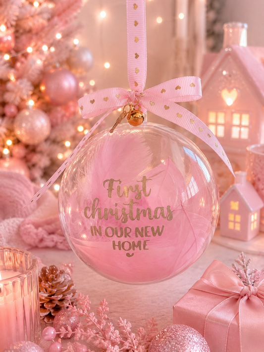 A clear round Christmas ornament with a pink inner layer and gold lettering that reads 'First Christmas IN OUR NEW HOME,' hanging from a pink ribbon with small gold hearts. The ornament is styled in a warm, festive setting with pink lights, a decorated tree, small house lanterns, and gift boxes.