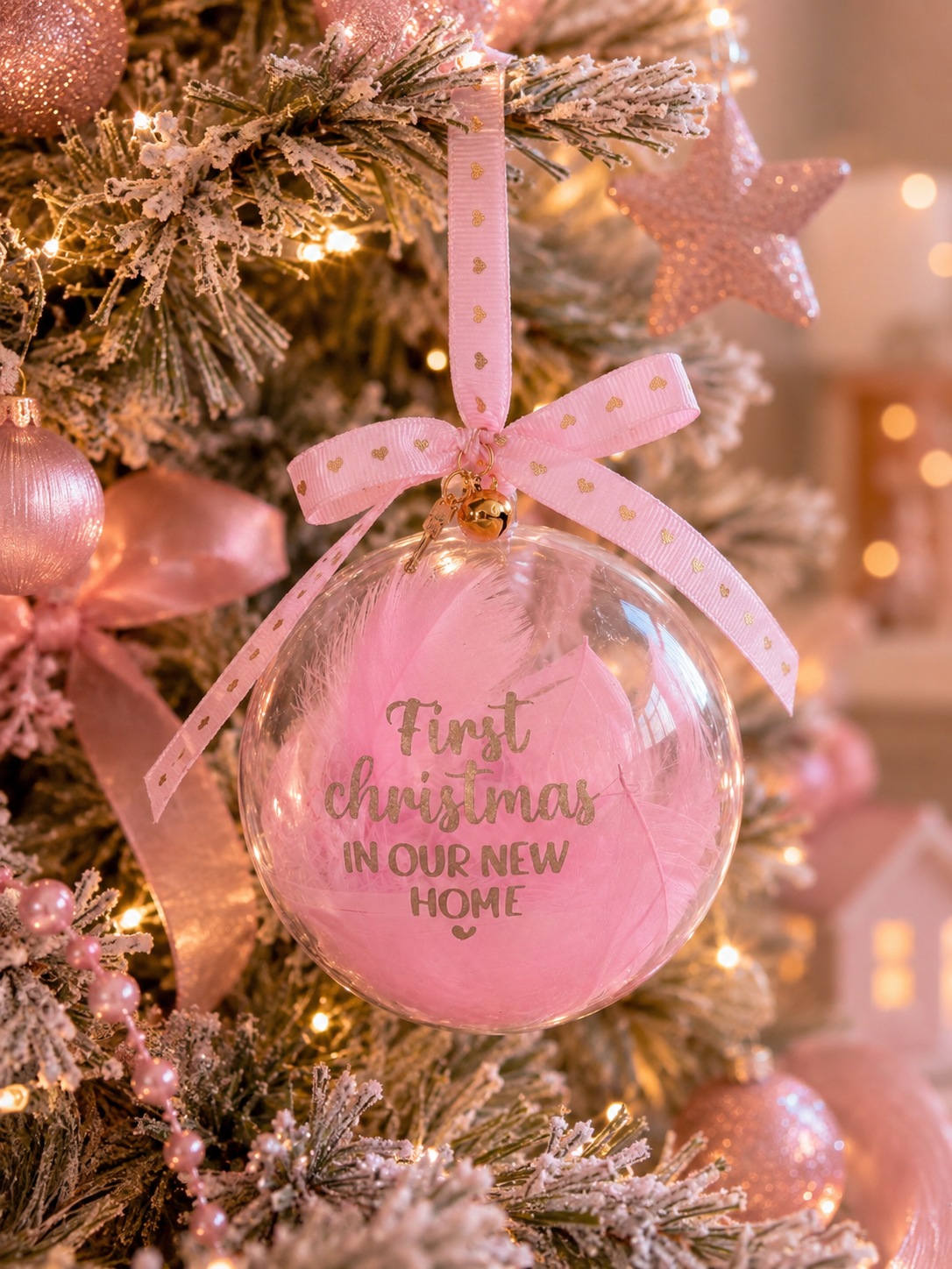 A round clear Christmas ornament with pink feather accents and gold foil text reading 'First Christmas IN OUR NEW HOME,' hanging on a decorated tree with pink ribbons and warm lights.