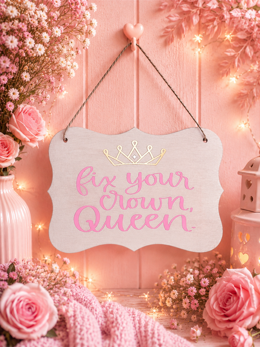 A decorative hanging sign with the phrase 'fix your crown, queen' in pink script and a small gold crown graphic at the top, displayed against a pink wooden wall with flowers, string lights, and pink textiles around it.