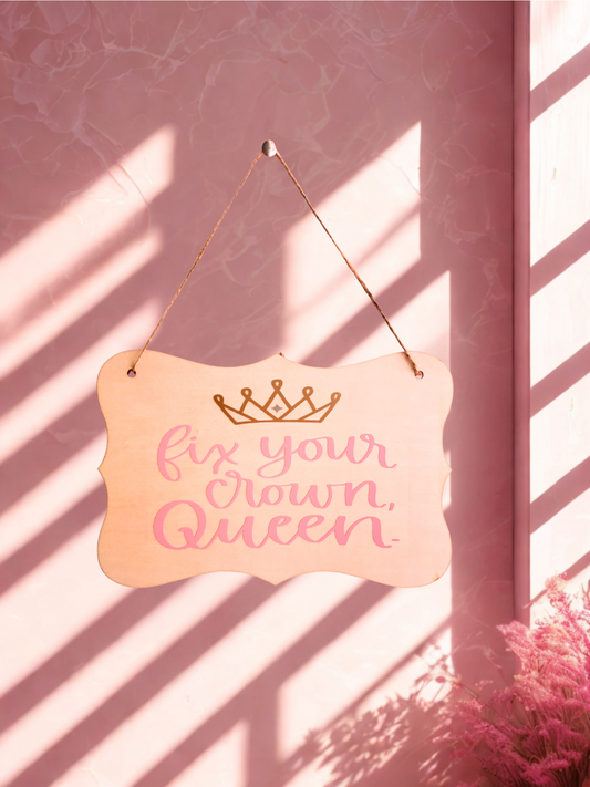 Decorative sign with 'Fix Your Crown Queen' text on a pink wall with sunlight casting shadows.