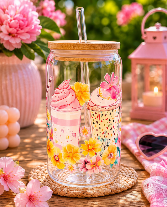 Glass tumbler with cupcake design and flowers on a wooden table with pink flowers and lantern in the background.