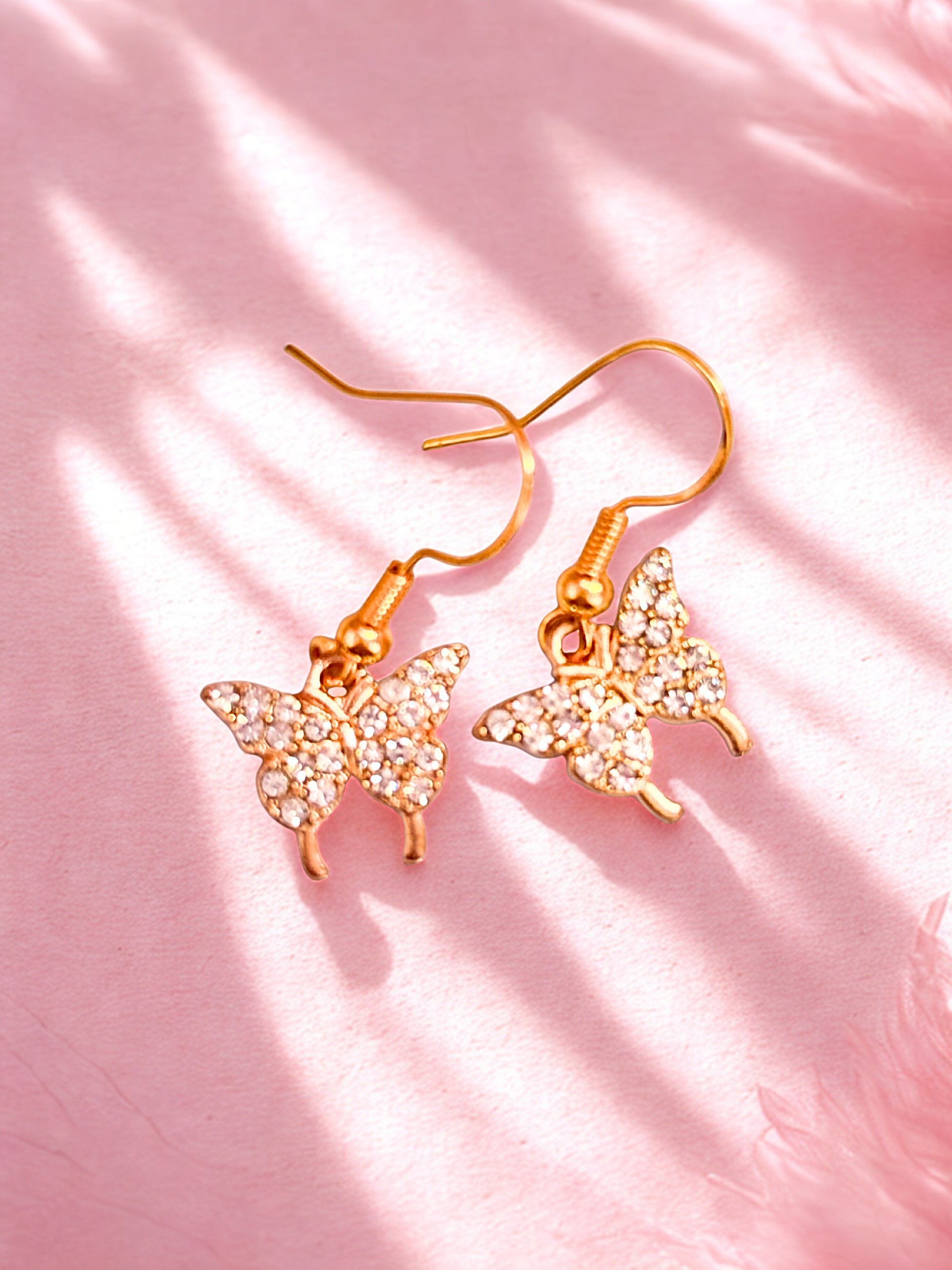 Butterfly-shaped earrings with gold and crystal details on a pink background