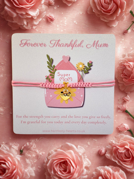 Pink branded Harmony Hearts card with 'Super Mom' and a pink cord bracelet with heart charm on a pink floral background