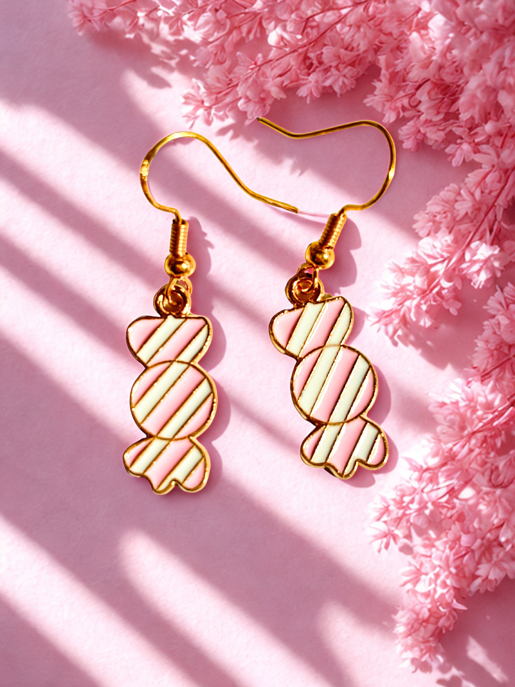 Gold pink and white candy-shaped sweet earrings on a pink background with pink flowers.