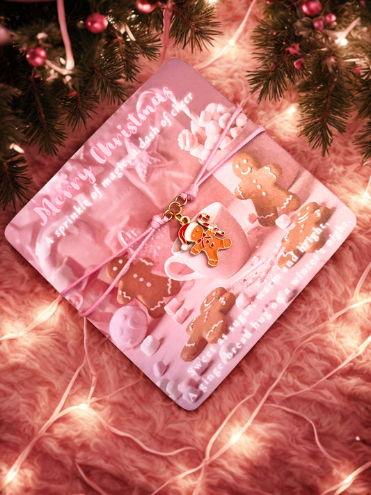 Pink Christmas-themed card with gingerbread cookies and a pink bow  and a gingerbread man charm bracelet on a soft pink background with lights.
