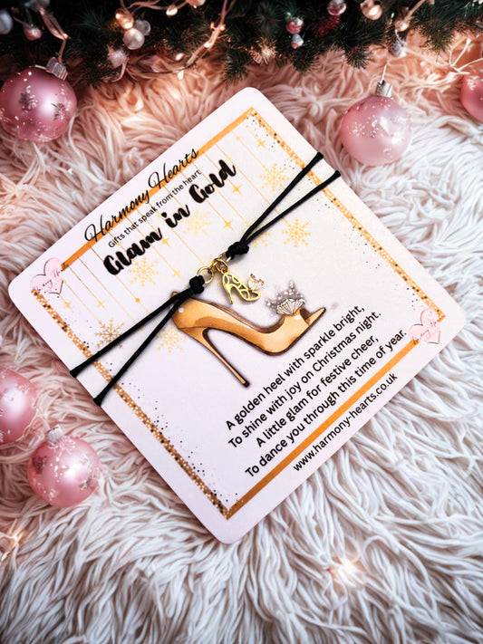 Gold shoe charm on a black cord bracelet with a card on a fluffy surface with Christmas decorations.