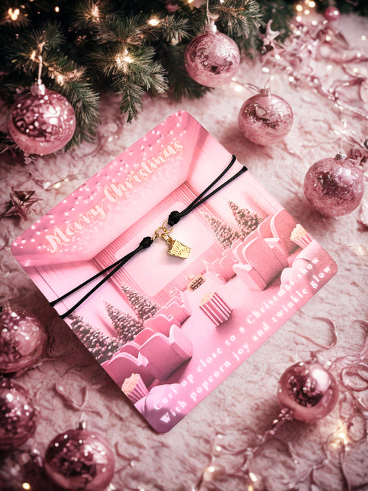 Pink Christmas-themed card with a popcorn charm bracelet and decorative ribbons on a festive background with pink ornaments and lights.