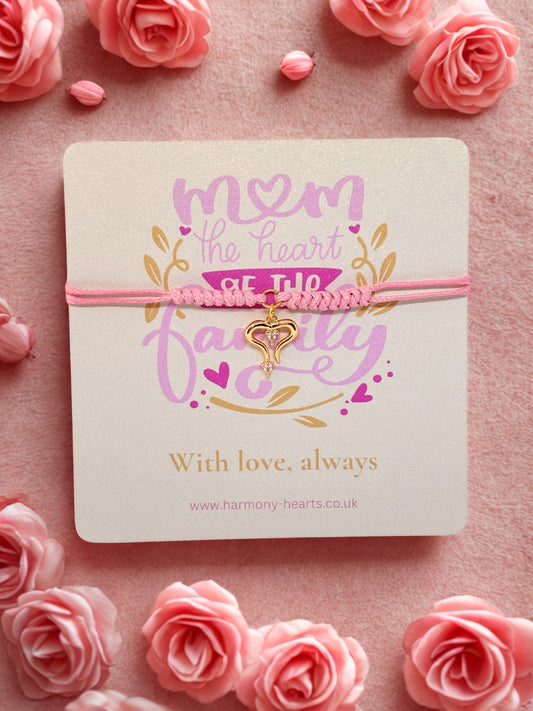 Pink bracelet with a sparkly gold heart charm presented on a Harmony Hearts branded card reading 'Mom, the heart of the family' on a pink floral background.