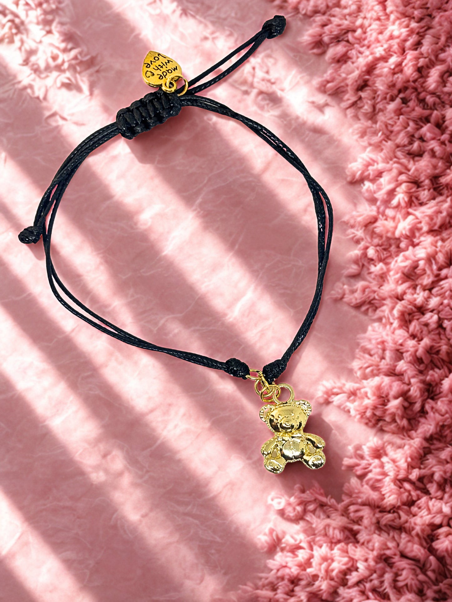 Black cord bracelet with gold teddy bear charm on a pink textured background