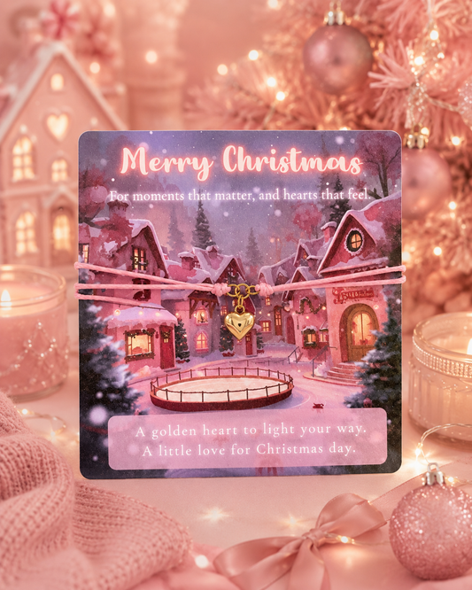 A square Christmas greeting card featuring a snowy village scene with houses, trees, and a small ice rink, tied with a pink cord bracelet and heart-shaped charm. The card reads 'Merry Christmas' and includes a quote at the bottom.