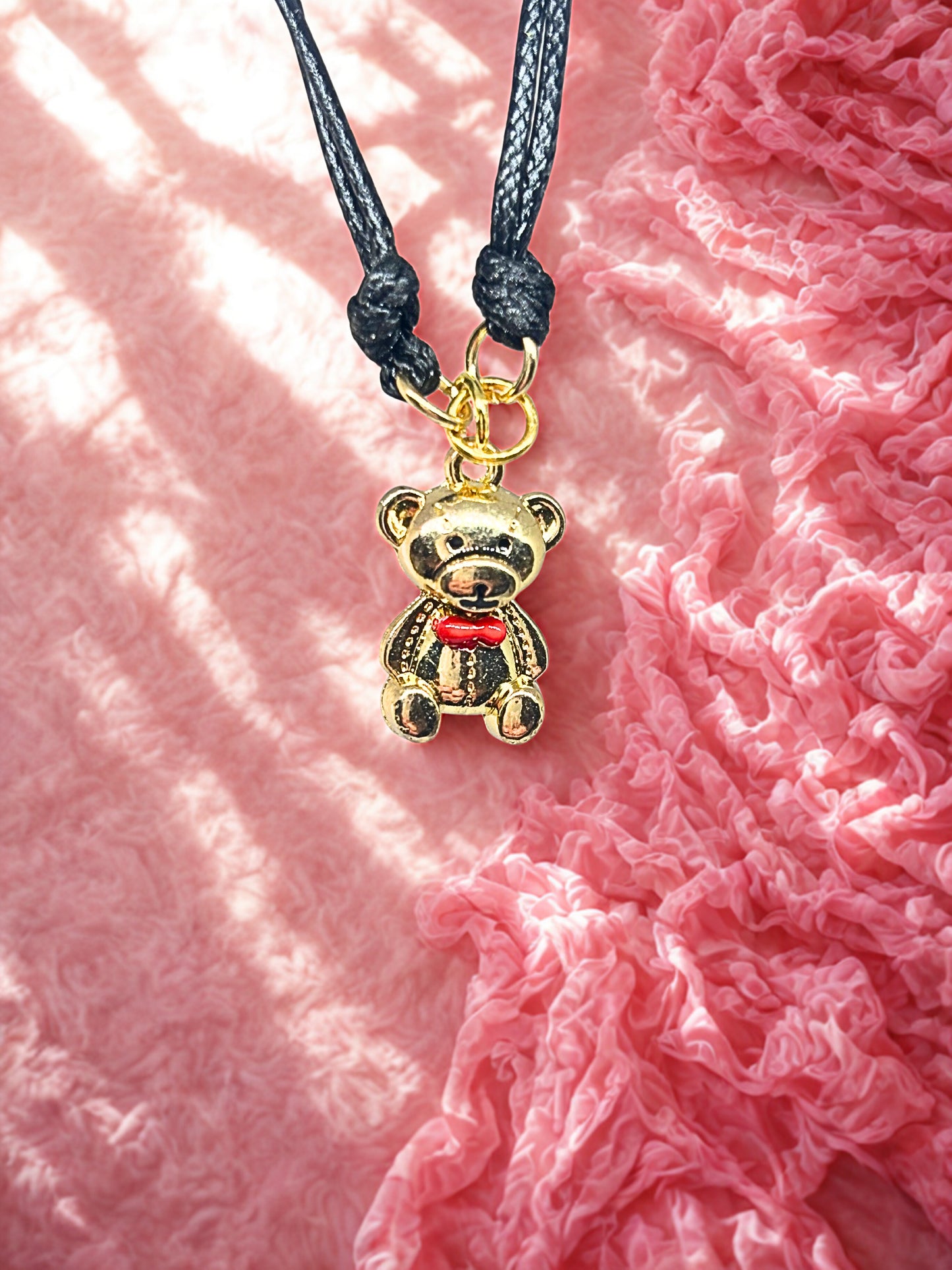 Gold teddy bear charm on a black cord against a pink textured background