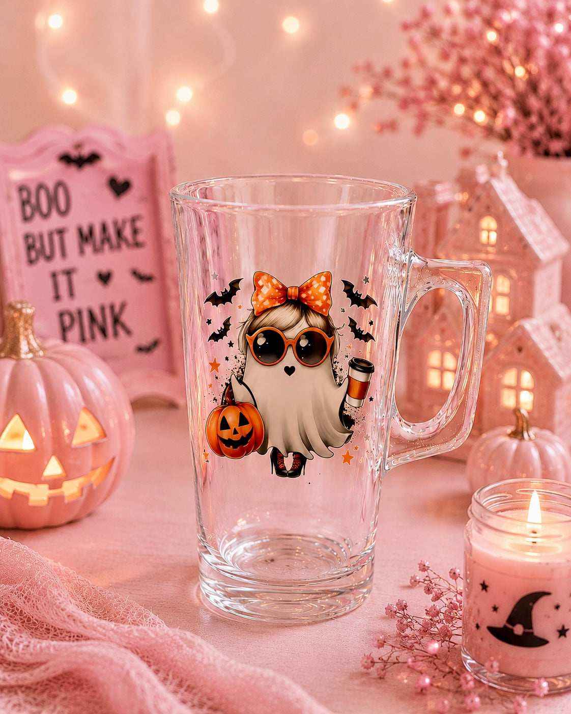 A clear glass mug with a Halloween-themed ghost illustration, set in a cozy, pink-lit tabletop scene with a jack-o'-lantern, candle, and decorative house.