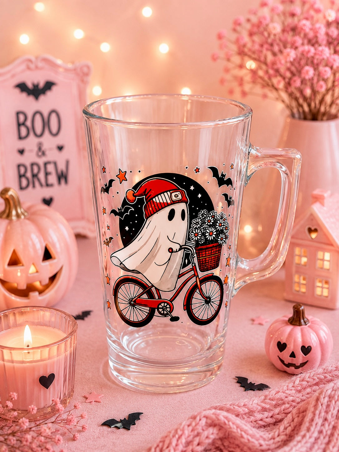 A clear glass mug with a Halloween-themed ghost and bicycle graphic, styled on a pink surface with pumpkins, a candle, a small house, and string lights in the background.