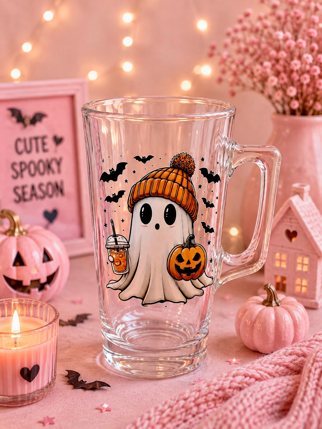 A clear glass mug with a Halloween-themed illustration of a ghost in an orange beanie holding a pumpkin and drink, surrounded by pink decorative props, string lights, and a candle.