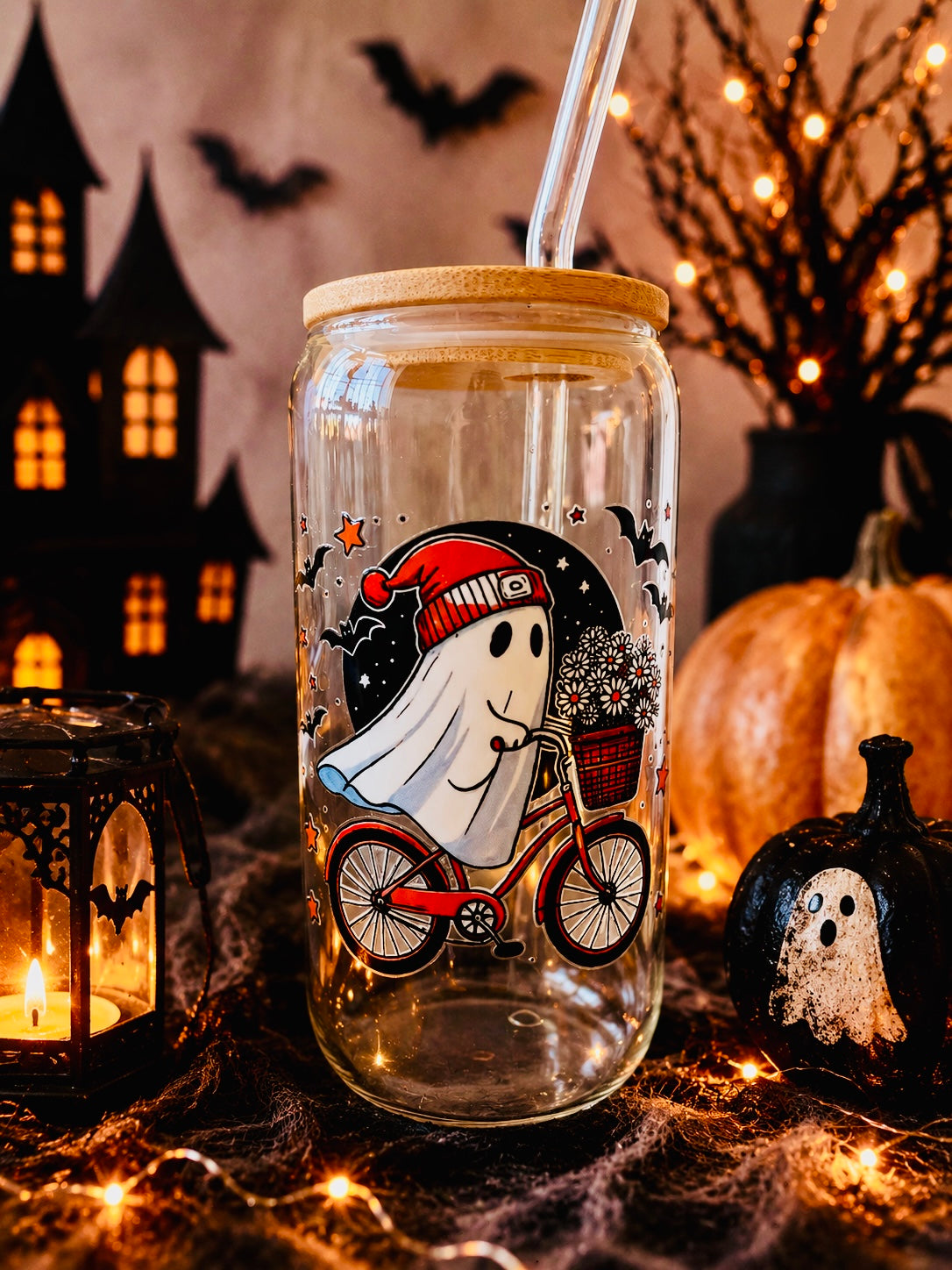 A clear glass mason jar tumbler with a bamboo lid and clear straw, decorated with a cartoon ghost riding a red bicycle. The jar is set in a cozy Halloween scene with a lit candle lantern, a ghost pumpkin, a small pumpkin, and warm fairy lights.