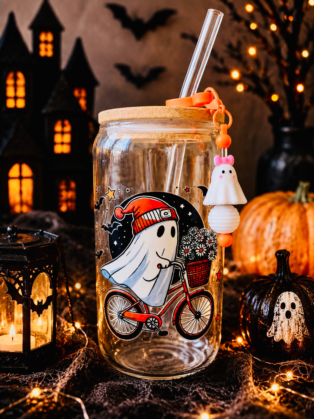 A clear glass mason jar with a bamboo lid and clear straw, decorated with a cartoon ghost riding a red bicycle on the front. The jar is styled in a Halloween setting with a ghost charm hanging from the straw, surrounded by a lantern, a small pumpkin, cobwebs, and a dark haunted house backdrop.
