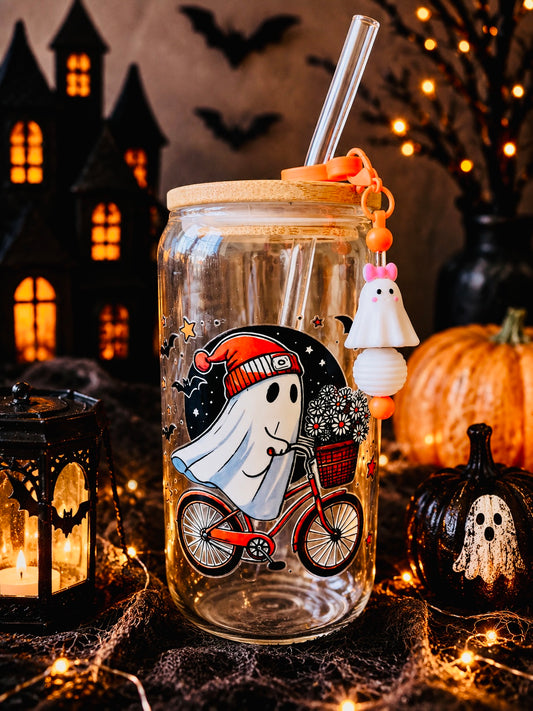 A clear glass mason jar with a bamboo lid and clear straw, decorated with a cartoon ghost riding a red bicycle on the front. The jar is styled in a Halloween setting with a ghost charm hanging from the straw, surrounded by a lantern, a small pumpkin, cobwebs, and a dark haunted house backdrop.