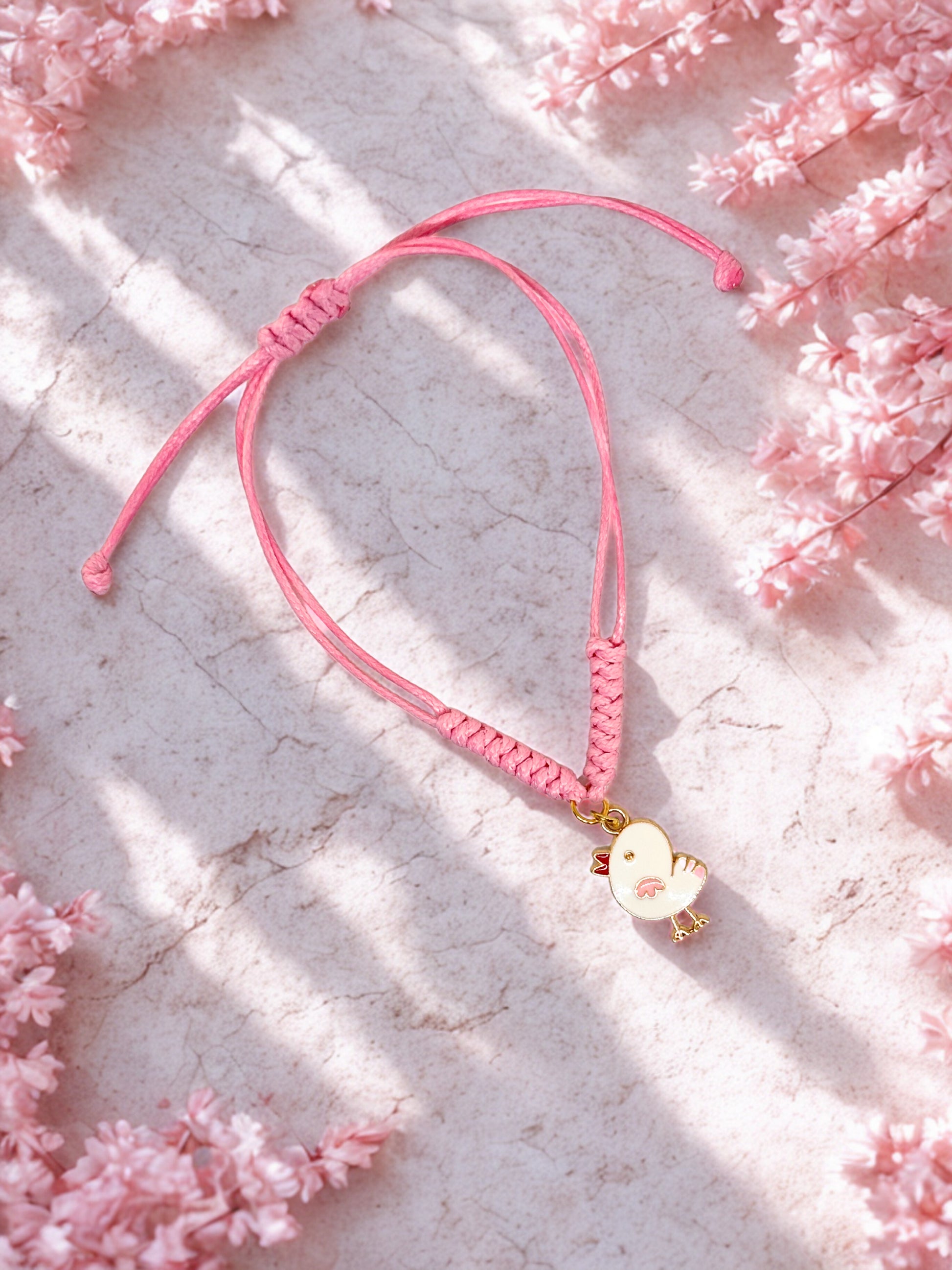 Pink bracelet with a chick  charm on a light stone surface with pink flowers.