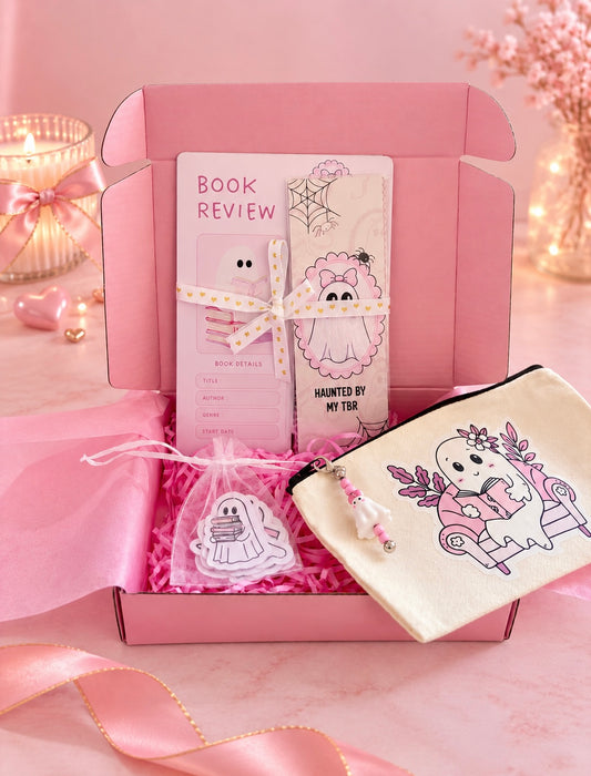 A pink gift box filled with cute stationery and accessories, including a book review card, bookmarks, a small bag with ghost stickers, and a cream pouch with a pink ghost illustration, arranged on a pink surface with soft decorative lights in the background.