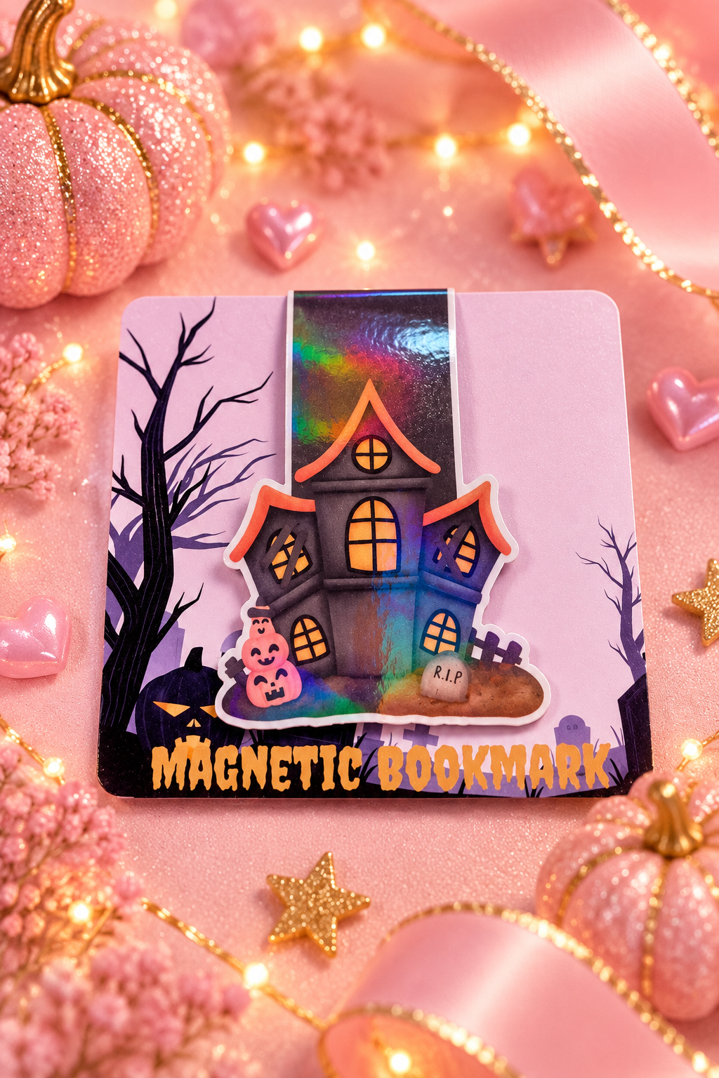 A Halloween-themed magnetic bookmark featuring a spooky haunted house, bare trees, pumpkins, and the text 'MAGNETIC BOOKMARK' on a pink backing card, styled with pink ribbons, glitter pumpkins, and warm fairy lights.