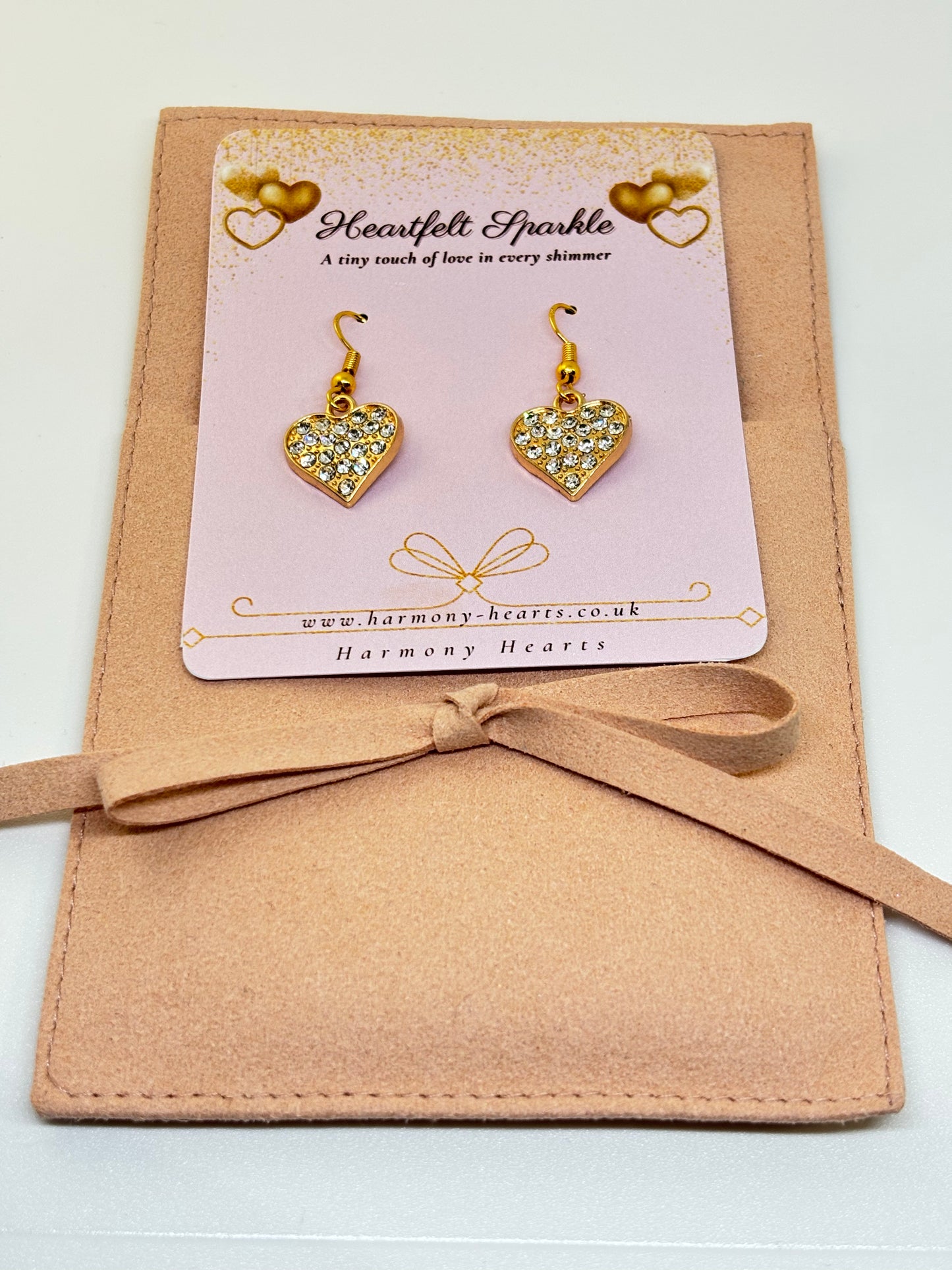 Heart-shaped earrings with gold and clear stones on a pink card with 'Harmony Hearts' branding, placed on a beige fabric background.
