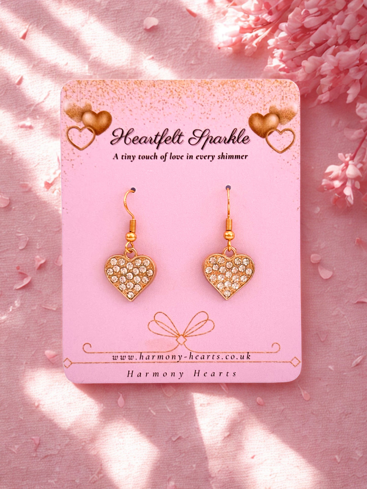 Heart-shaped earrings on a pink card with 'Harmony Hearts' branding, set against a pink floral background.