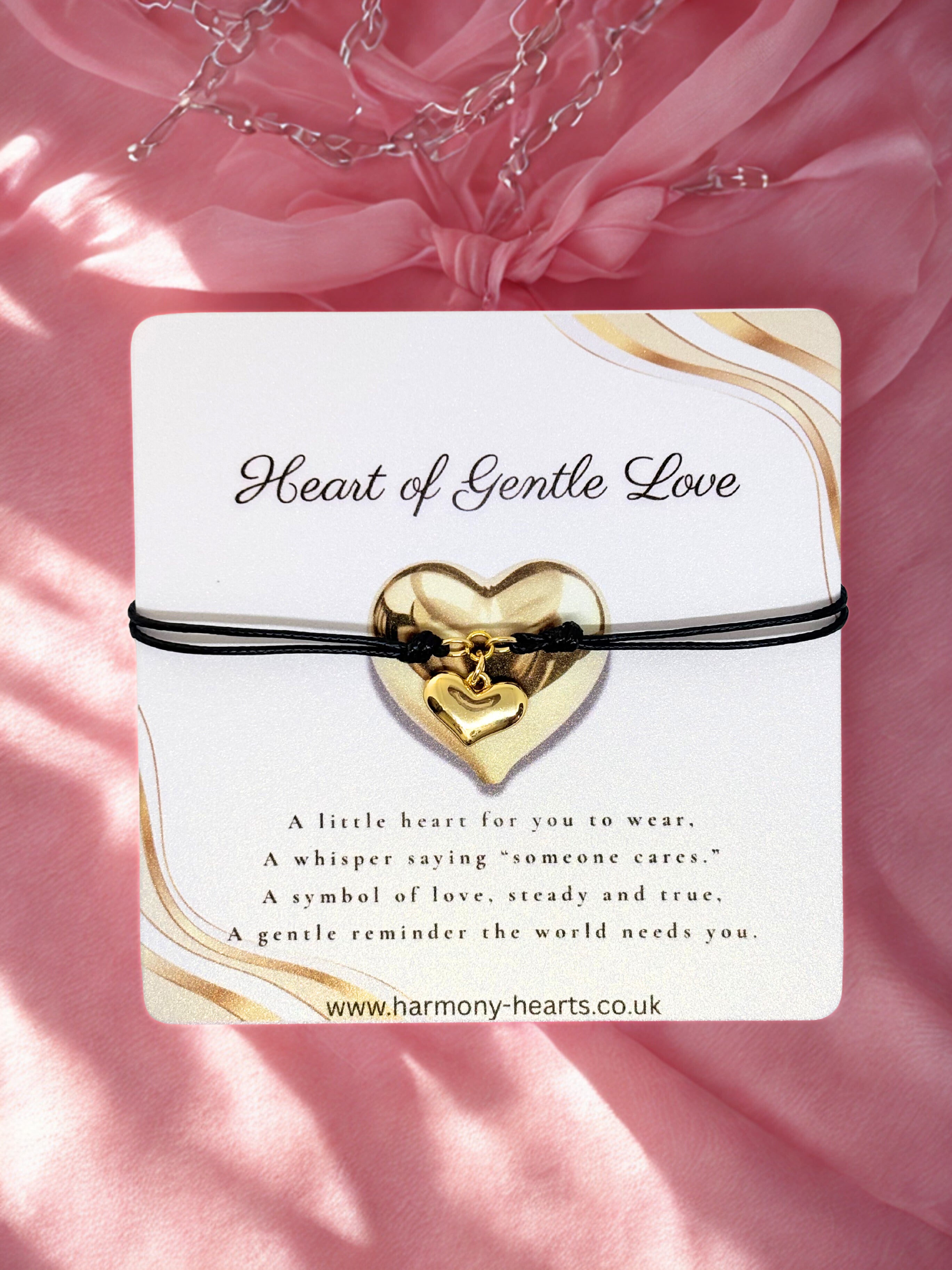 Gold heart-shaped bracelet on a card with 'Heart of Gentle Love' text against a pink fabric background.
