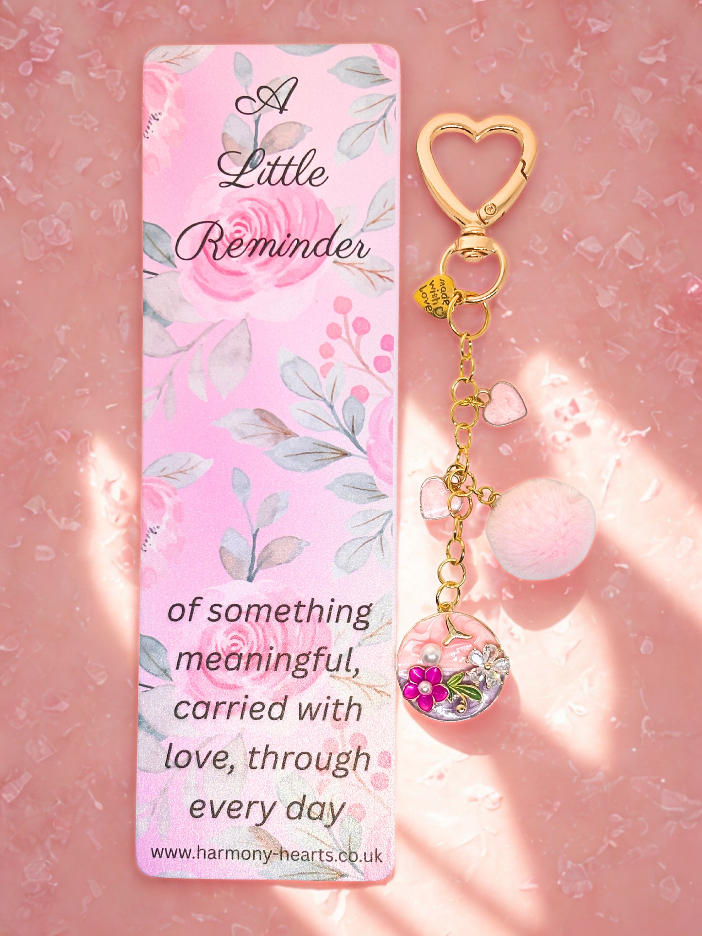 Keychain bag charm with heart design and floral charm on a pink card with text, against a pink background.