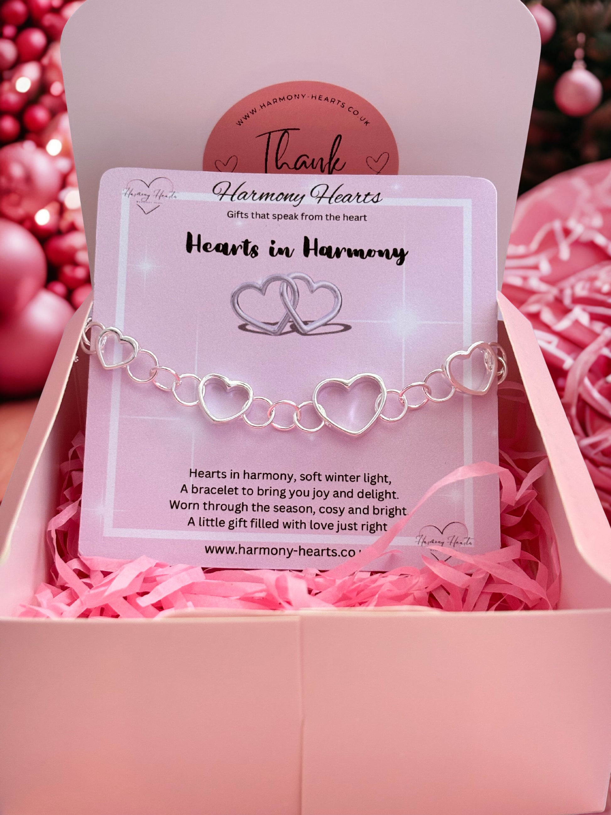 Silver bracelet with heart design on a card in a pink box, surrounded by pink decorative elements.