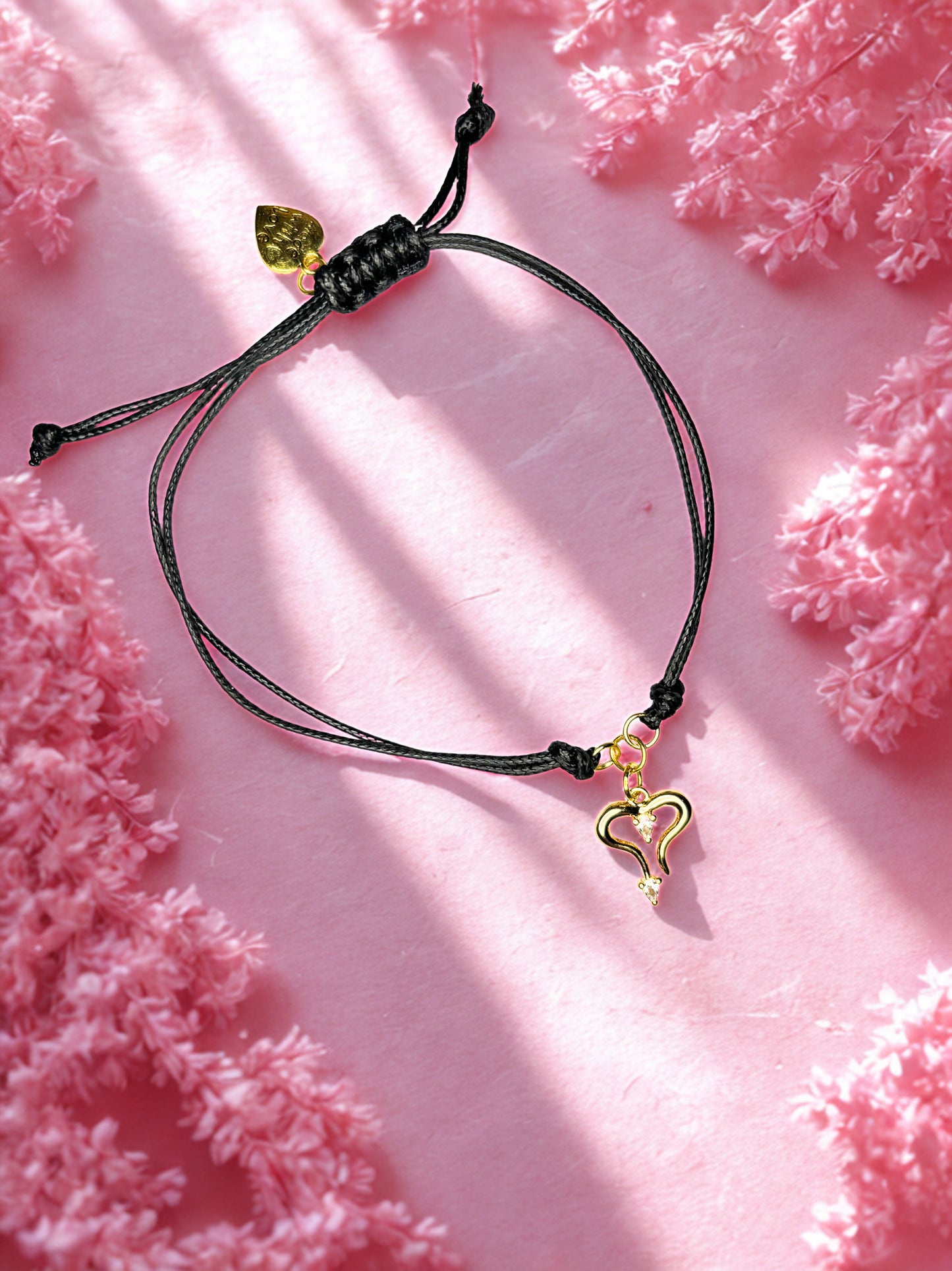 Black cord bracelet with gold heart charm on a pink textured background