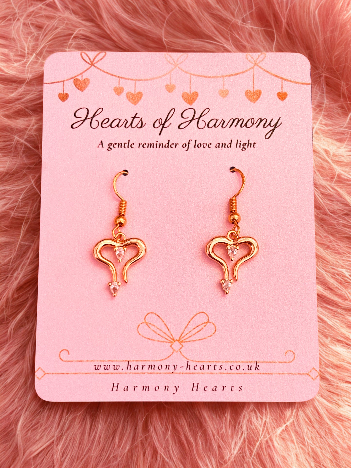 Heart-shaped earrings on a 'Hearts of Harmony' branded card against a pink fluffy background