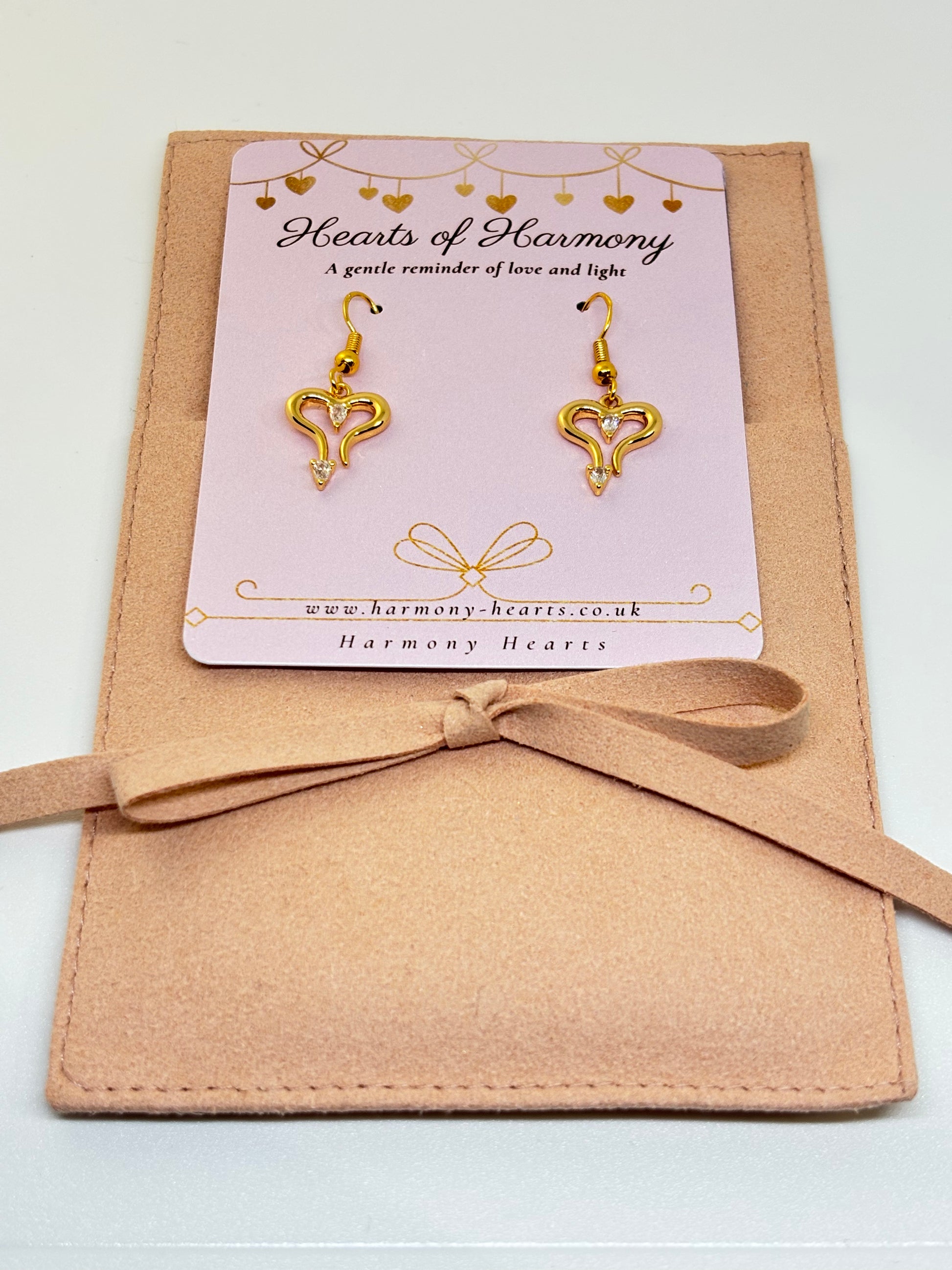 Gold heart-shaped earrings on a 'Hearts of Harmony' card with a beige ribbon.