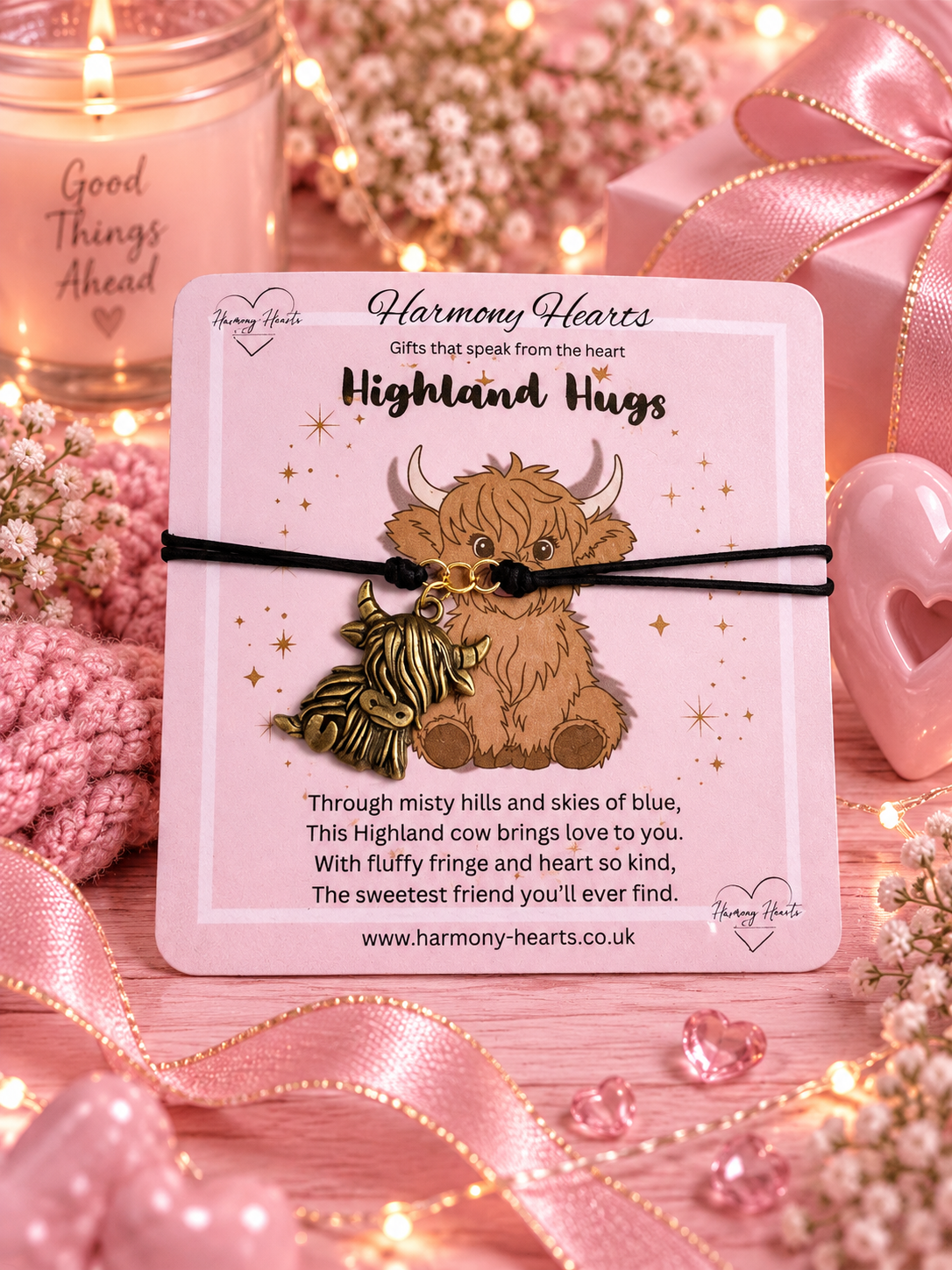 A handmade friendship bracelet with a Highland cow and charm, presented on a branded pink card labeled 'Harmony Hearts' and 'Highland Hugs'. The card is styled in a cozy, romantic setting with pink ribbons, a lit candle, and fairy lights.
