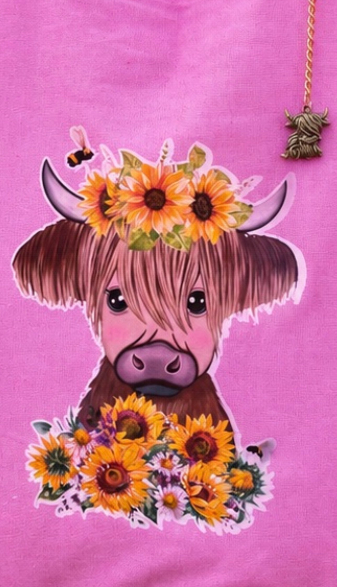 A close-up of a pink tote bag featuring a cartoon Highland cow graphic with a sunflower crown, flowers at the bottom, and a bee near the top right.