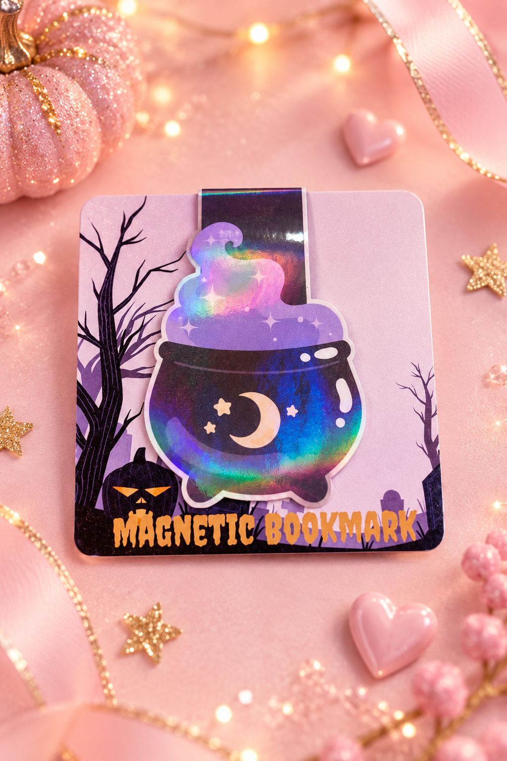 A Halloween-themed magnetic bookmark featuring a colorful cauldron with a galaxy night sky design, surrounded by spooky trees and a jack-o'-lantern, with the text 'MAGNETIC BOOKMARK' at the bottom. The bookmark is displayed on a pink background decorated with a glittery pumpkin, heart-shaped beads, and warm fairy lights.