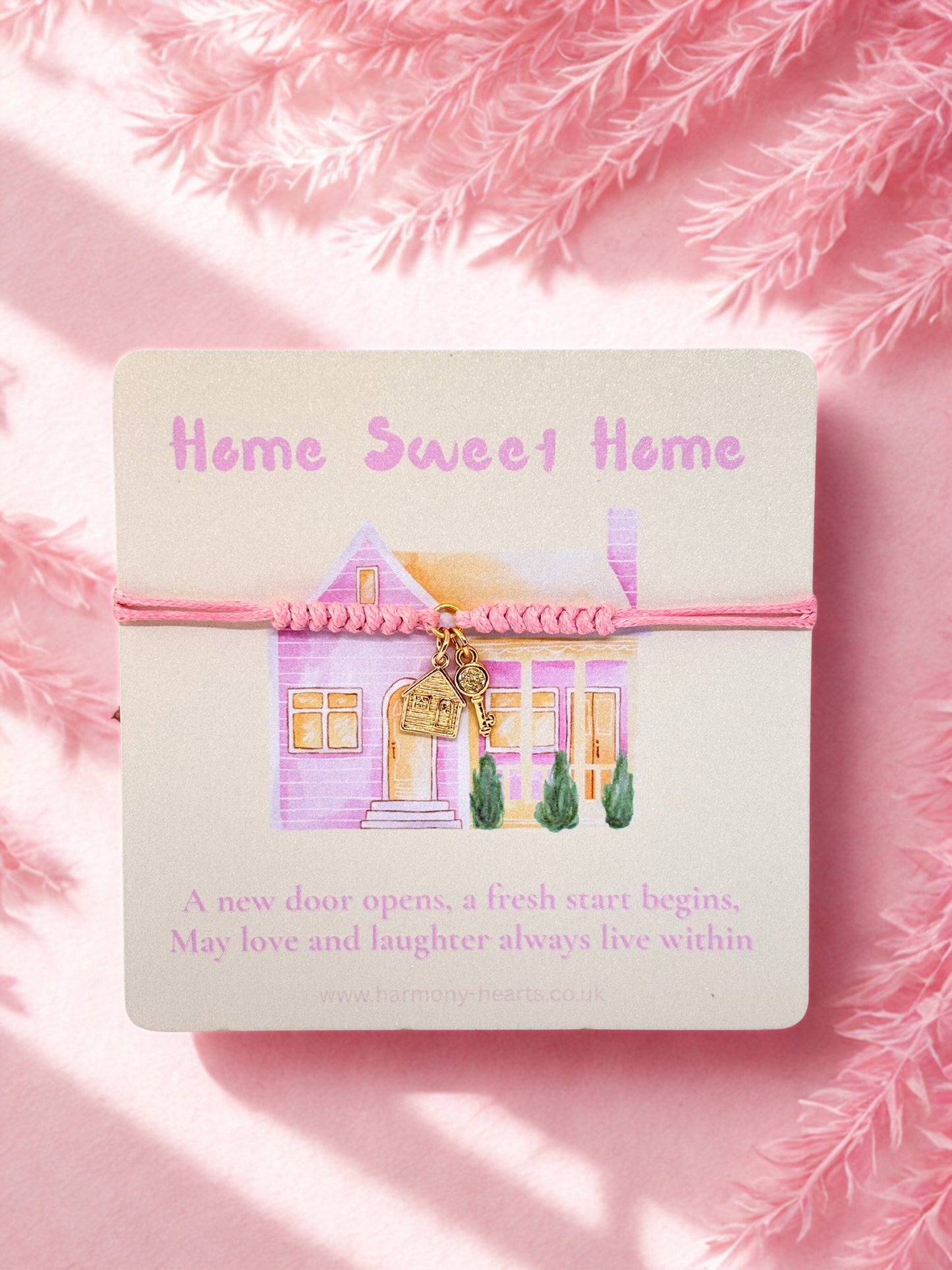 Pink cord bracelet with house and key charms on a card with 'Home Sweet Home' text on a pink background