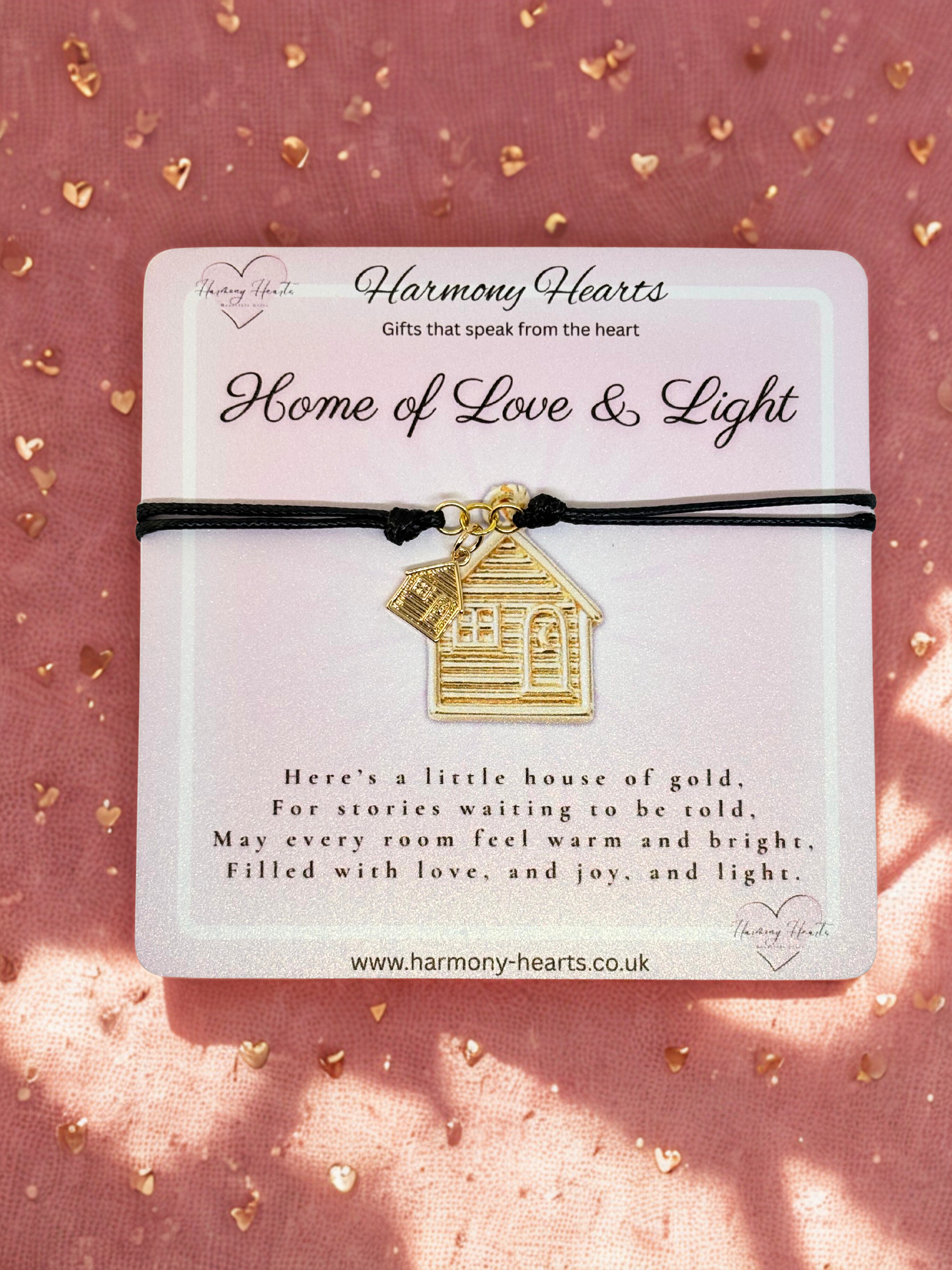 Gold house-shaped charm on a black cord with 'Harmony Hearts' packaging on a pink background.