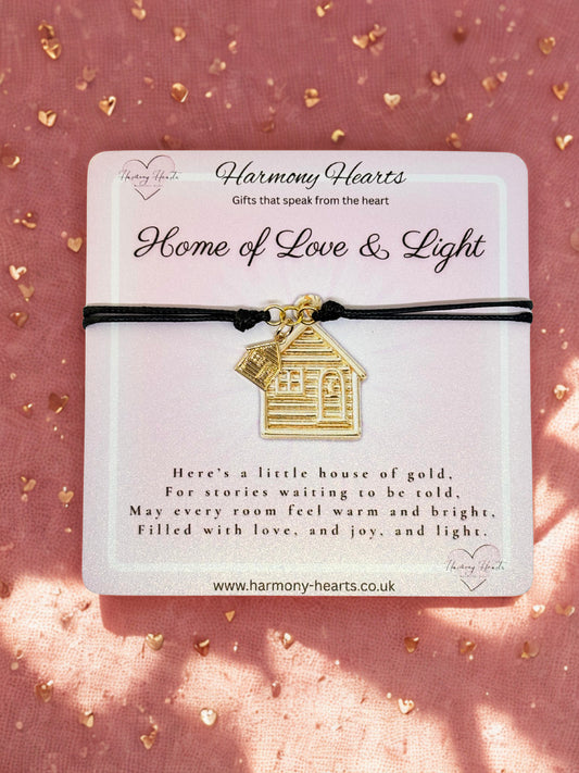 Gold house-shaped charm on a black cord with 'Harmony Hearts' packaging on a pink background.