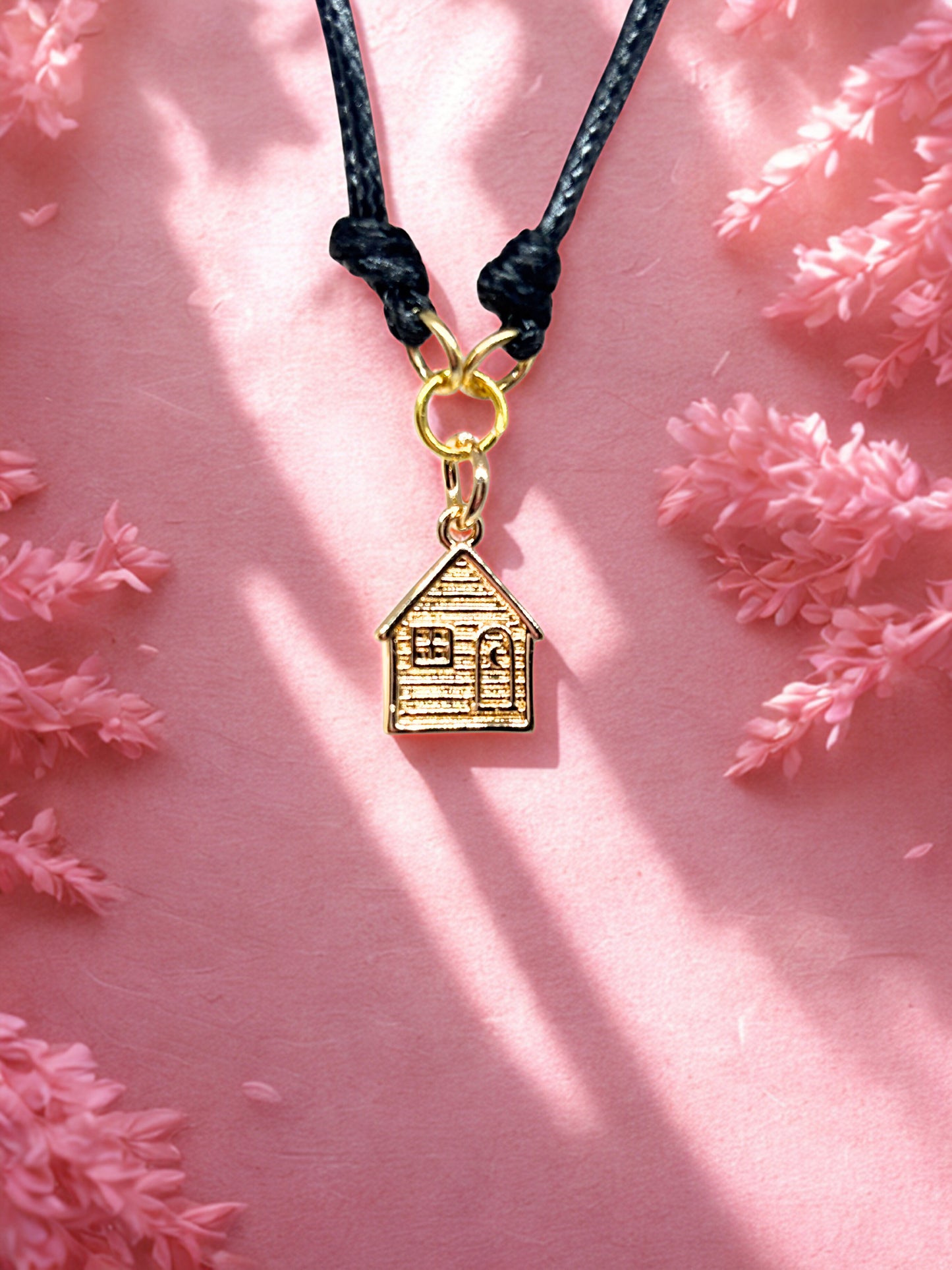Gold house-shaped charm on a black cord against a pink floral background