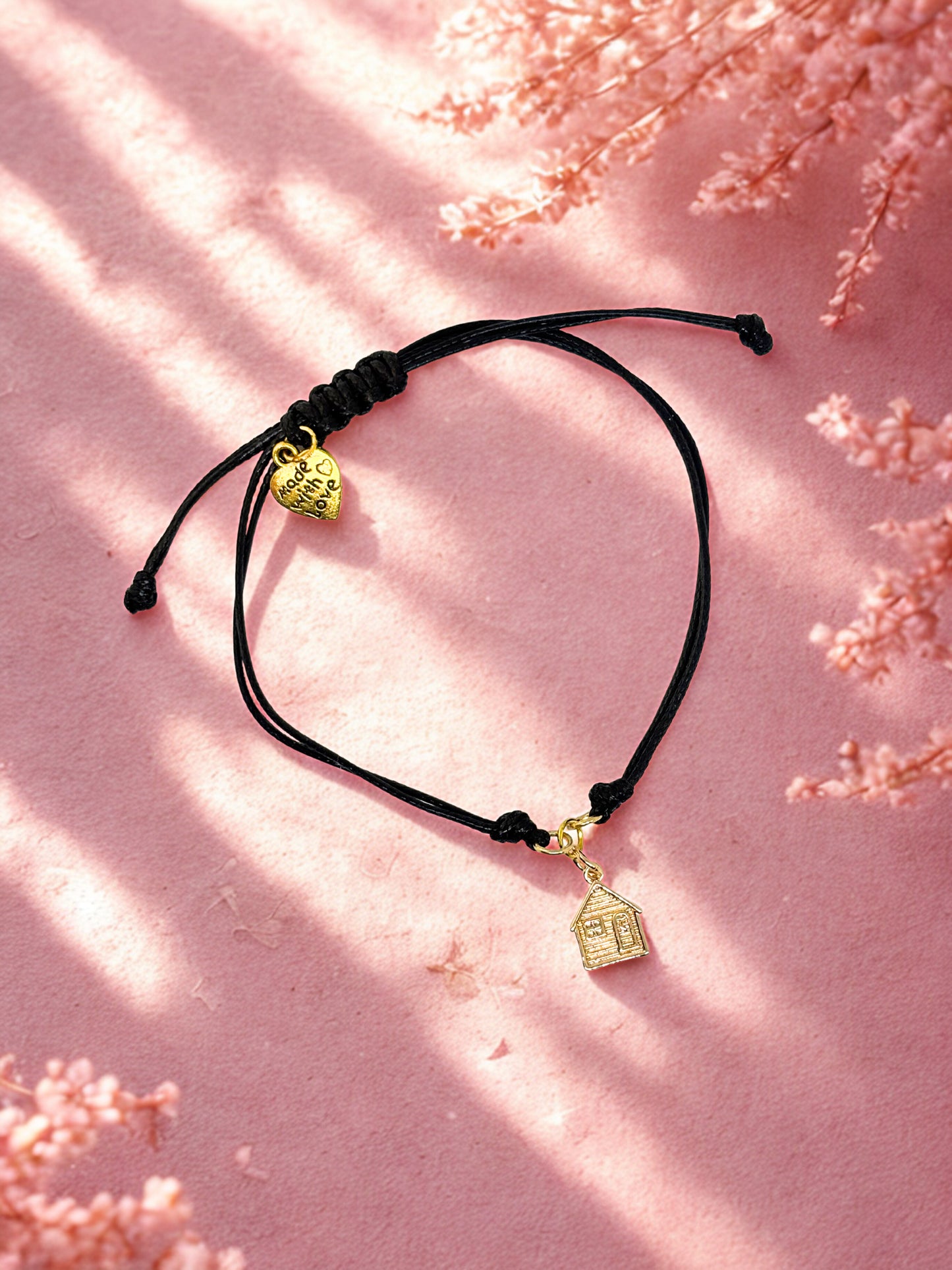 Black cord bracelet with gold house charm on a pink floral background