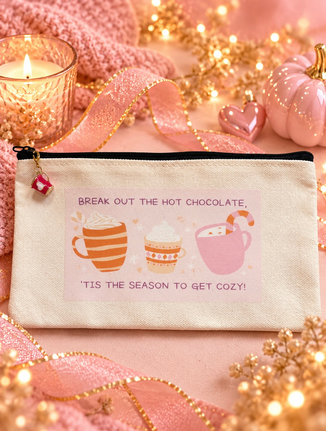 A canvas zip pouch with a cozy hot chocolate theme, featuring illustrations of three hot chocolate cups and the text 'BREAK OUT THE HOT CHOCOLATE, TIS THE SEASON TO GET COZY!'. The pouch is styled in a warm, festive setting with pink ribbons, a lit candle, and gold fairy lights.