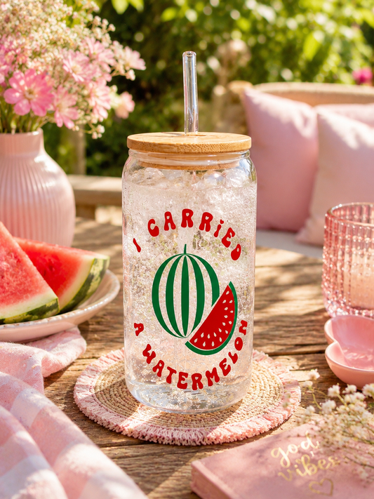 A clear glass tumbler with a bamboo lid and glass straw, printed with a watermelon graphic and the text 'I CARRIED A WATERMELON'. It is styled on a wooden table in a sunny, pink-themed outdoor setting with watermelon slices and pink accessories.