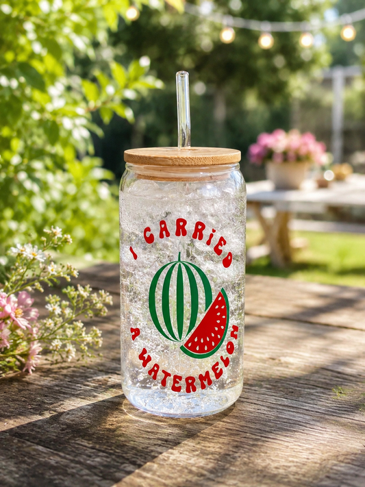 A clear glass tumbler with a bamboo lid and clear straw, featuring a watermelon graphic and the text 'I CARRIED A WATERMELON' in red, set on a wooden table in a sunny garden.