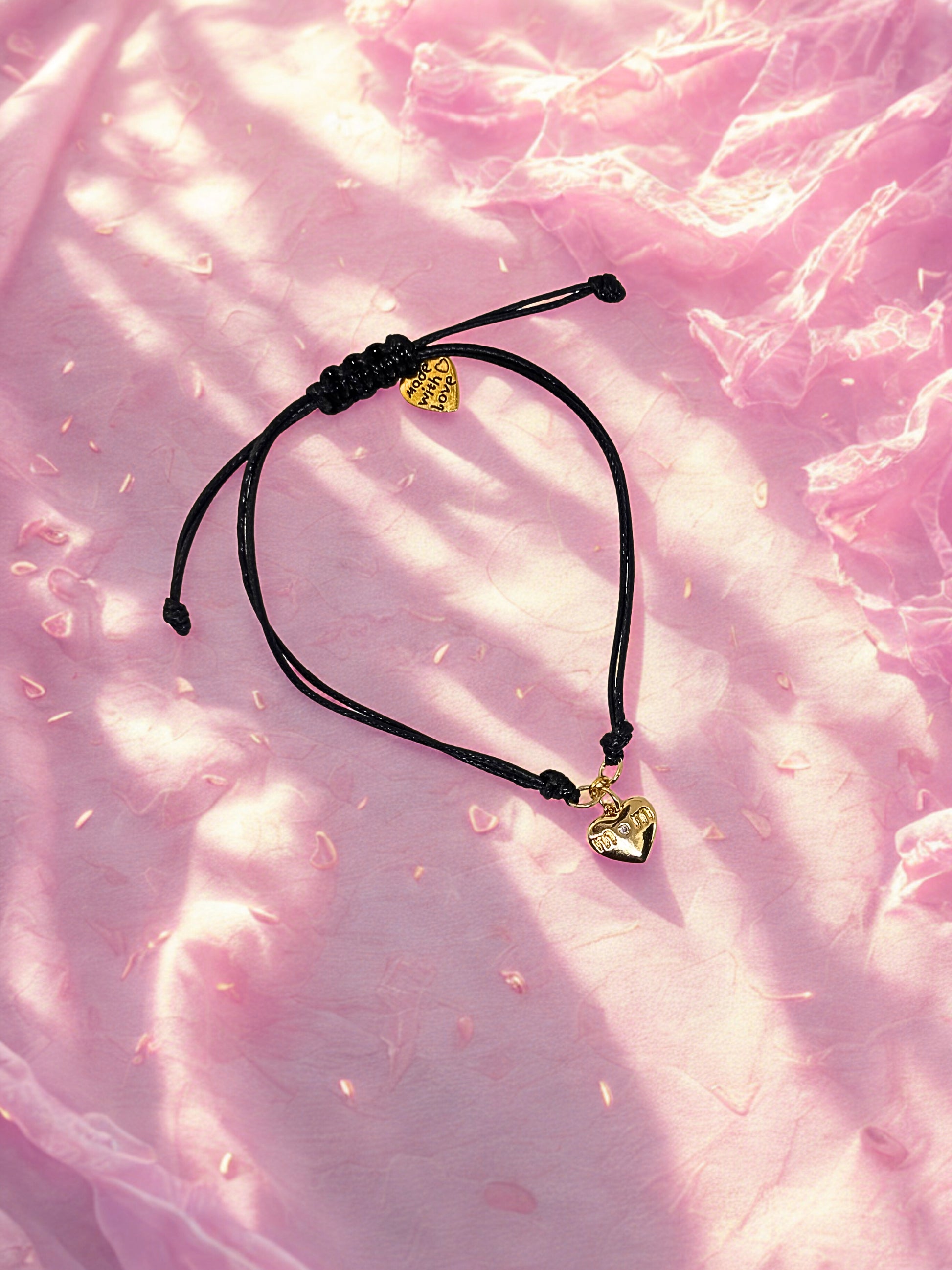 Black cord bracelet with gold heart charms on a pink textured background